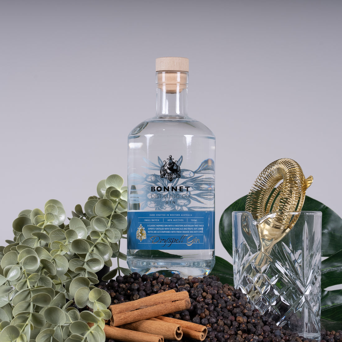 A bottle of DrySpell Gin surrounded by juniper berries, cinnamon sticks and eucalyptus, with a gold strainer and cut crystal glass